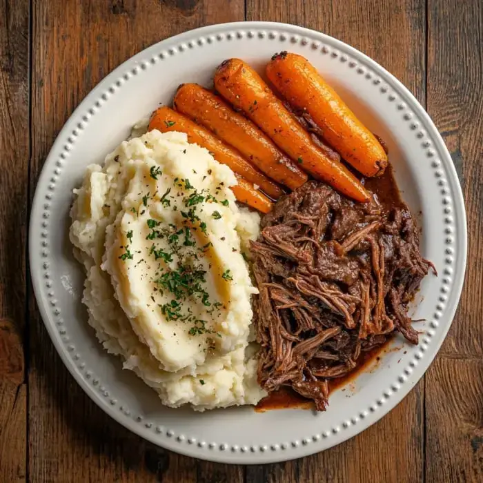 A plate of mashed potatoes topped with herbs, served alongside tender shredded beef and glazed carrots.