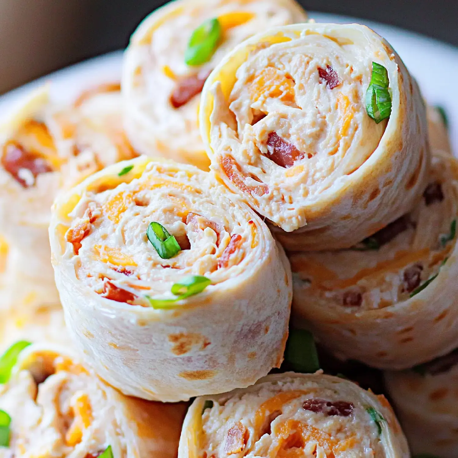 A close-up of rolled tortilla wraps filled with cheese, bacon, and green onions, stacked on a plate.