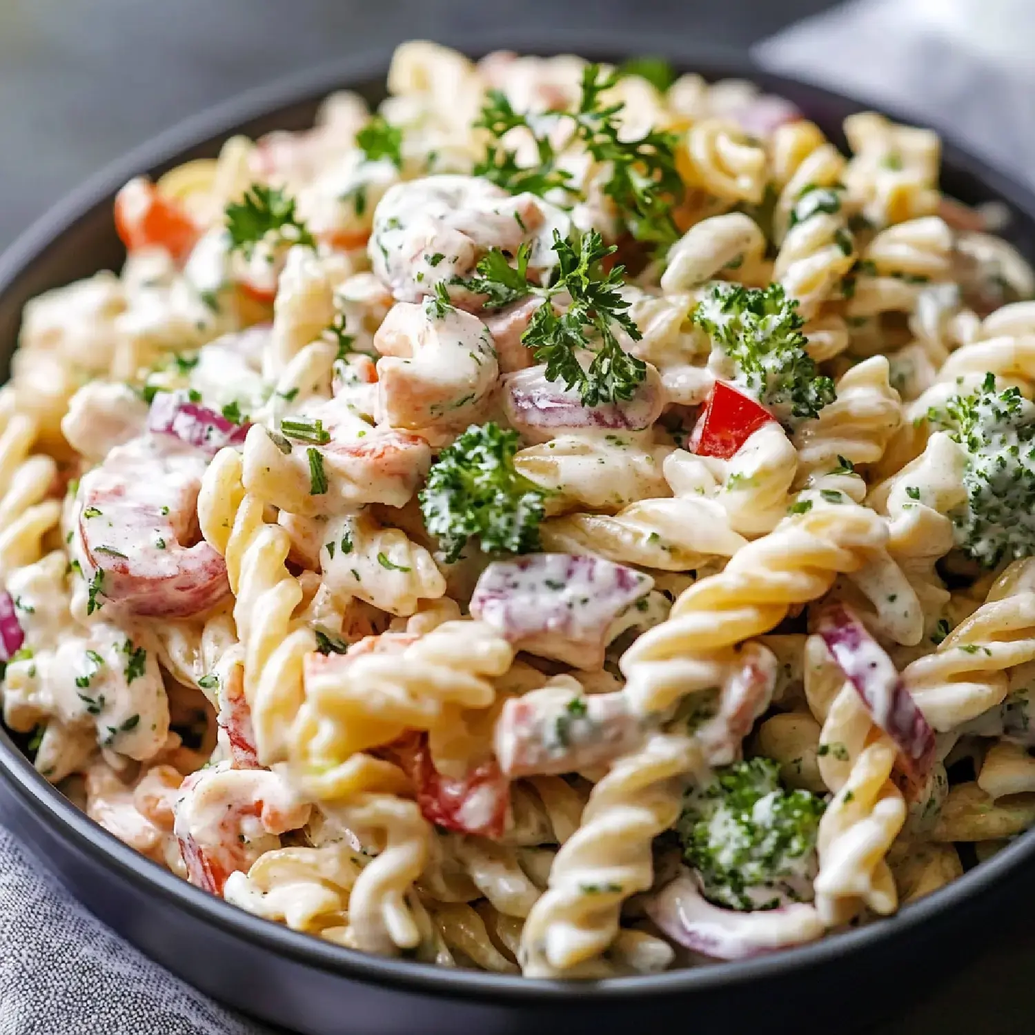 A close-up image of a bowl of creamy pasta salad featuring twisted pasta, mixed vegetables, and garnished with fresh parsley.