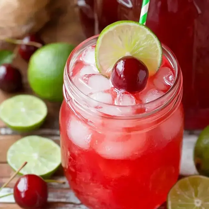 A refreshing red drink with ice, garnished with a lime slice and a cherry, in a mason jar, surrounded by lime halves and cherries.