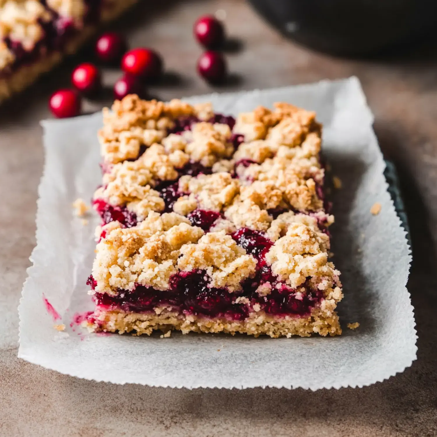 A slice of cranberry crumble dessert is displayed on a parchment-lined surface, with a few cranberries scattered in the background.