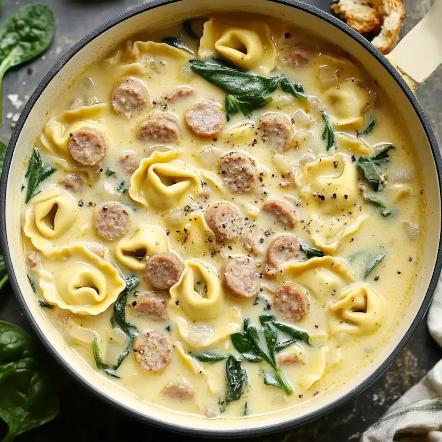 A creamy soup with tortellini, sausage, and spinach, garnished with black pepper, served in a pot.