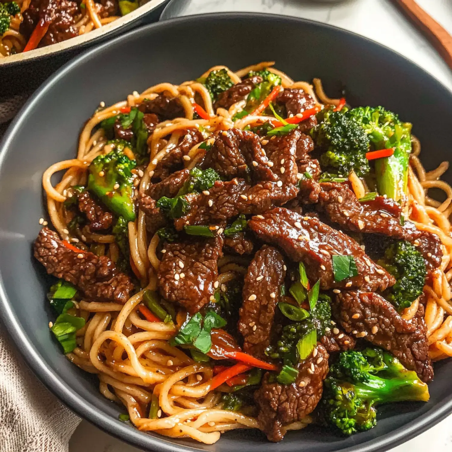 A close-up image of a bowl of beef stir-fry with broccoli and noodles, garnished with sesame seeds and red pepper strips.