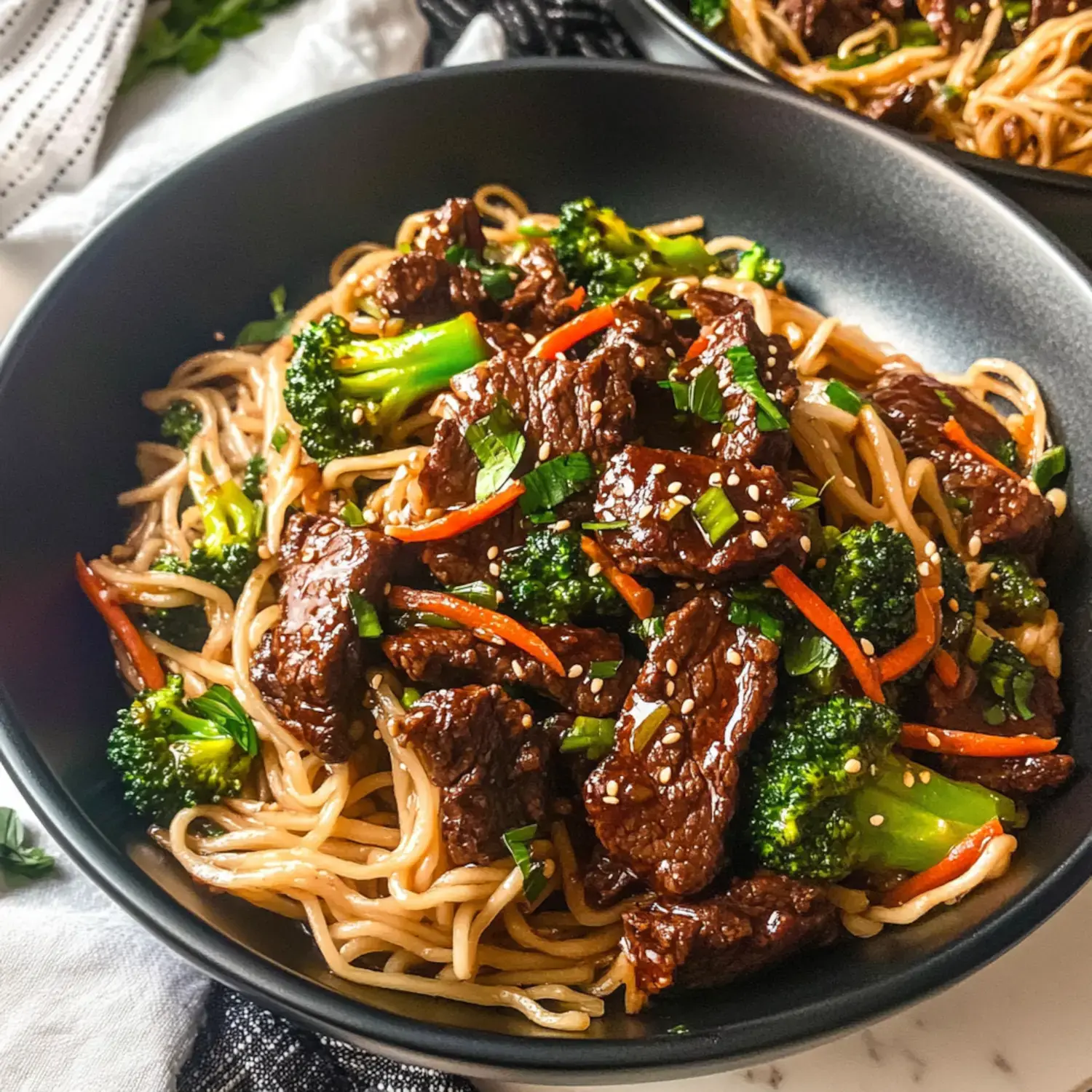 A close-up view of a bowl of stir-fried noodles with beef, broccoli, and carrots, garnished with green onions and sesame seeds.