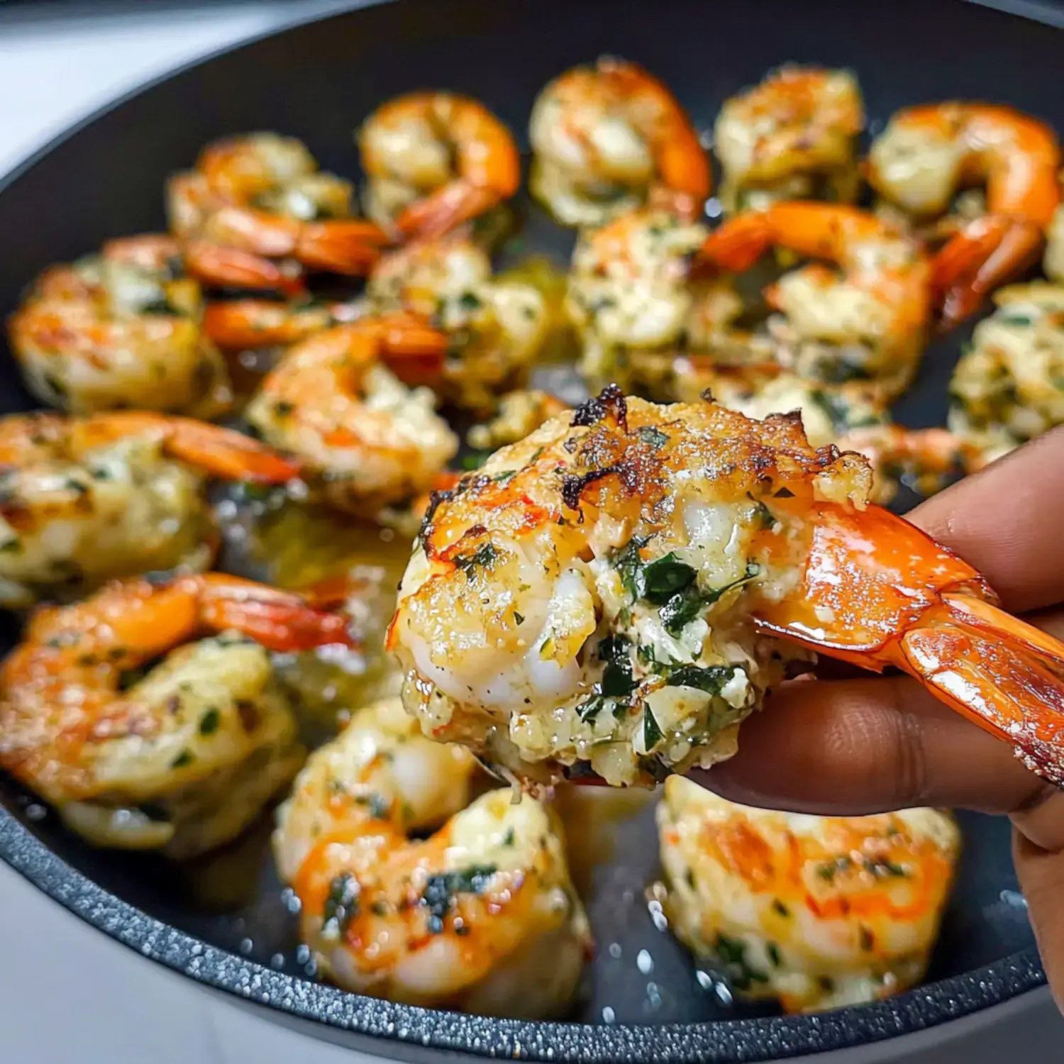 A hand holds a cooked shrimp filled with herbs and spices, surrounded by more shrimp in a frying pan.