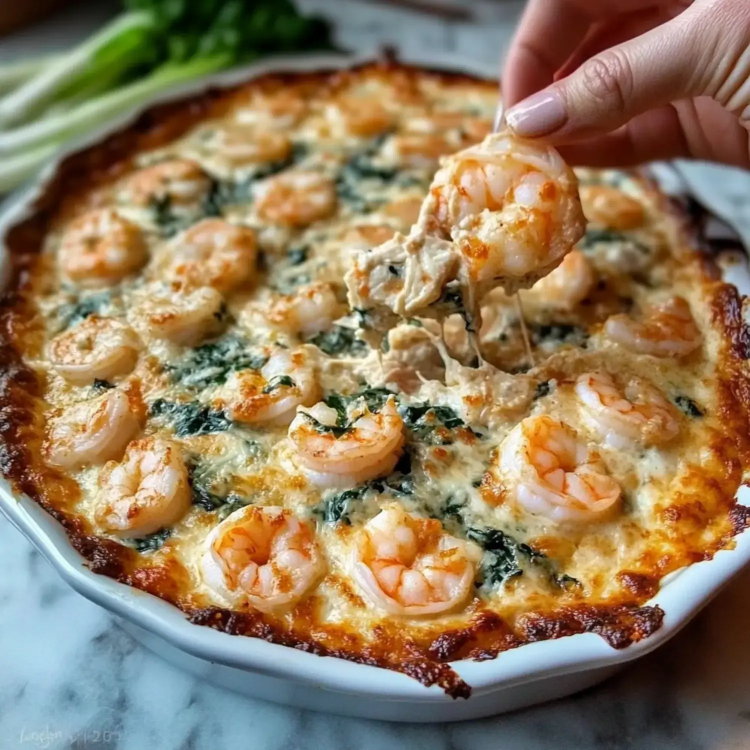 A person is lifting a cheesy, baked shrimp dish from a white serving dish, revealing shrimp and spinach in a creamy topping.