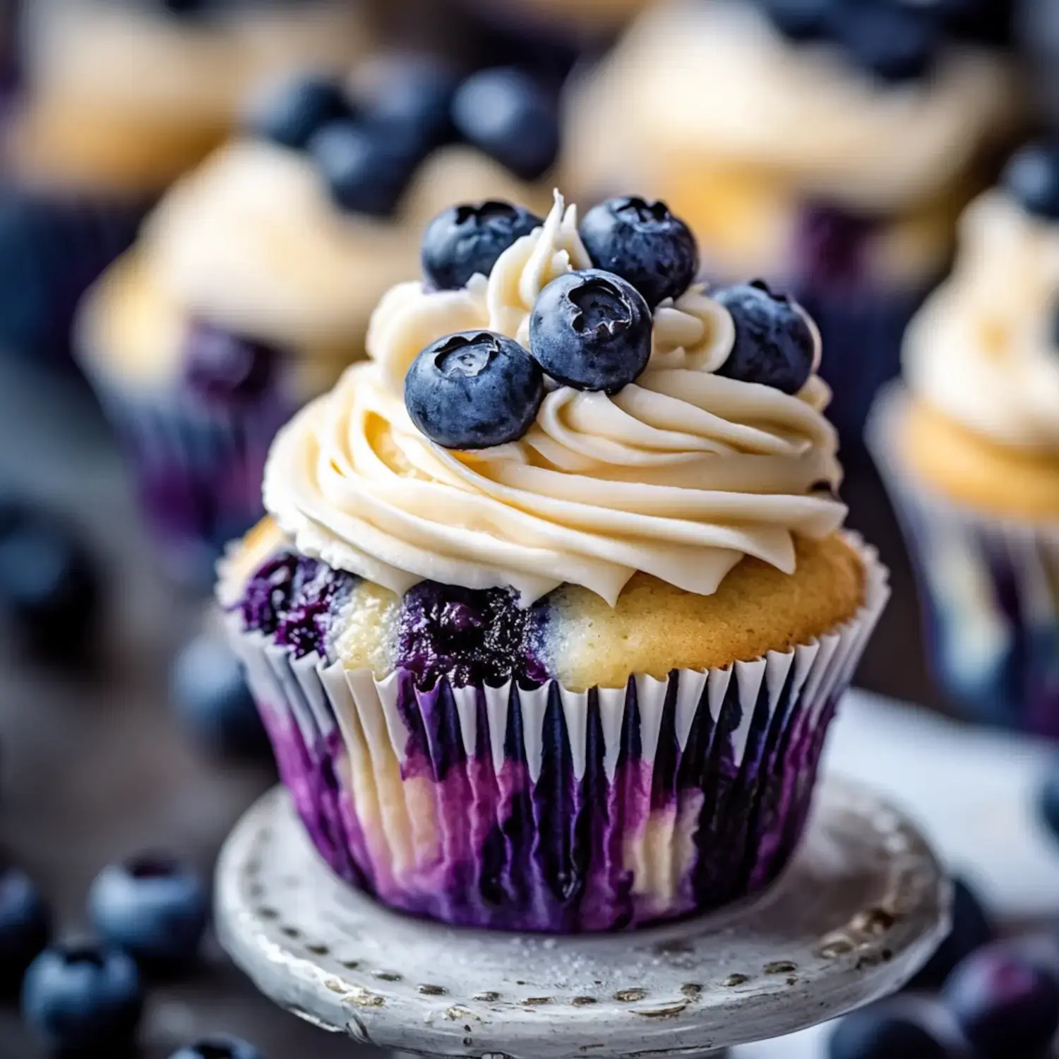 A close-up of a decorated blueberry cupcake topped with swirls of frosting and fresh blueberries, placed on a small stand.