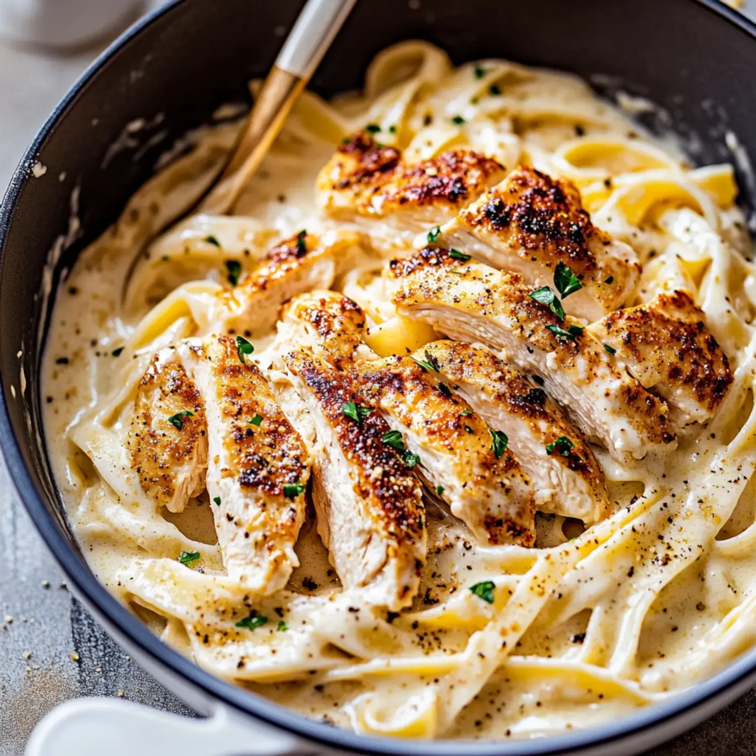 A close-up of creamy fettuccine pasta topped with slices of grilled chicken and garnished with parsley in a black skillet.