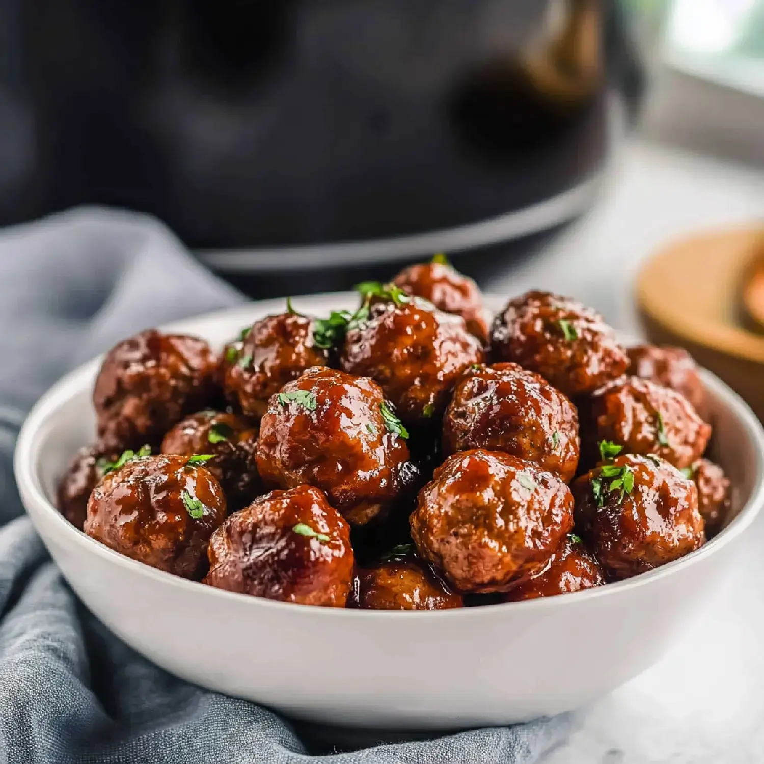 A bowl of glossy, glazed meatballs garnished with herbs, placed on a gray cloth.