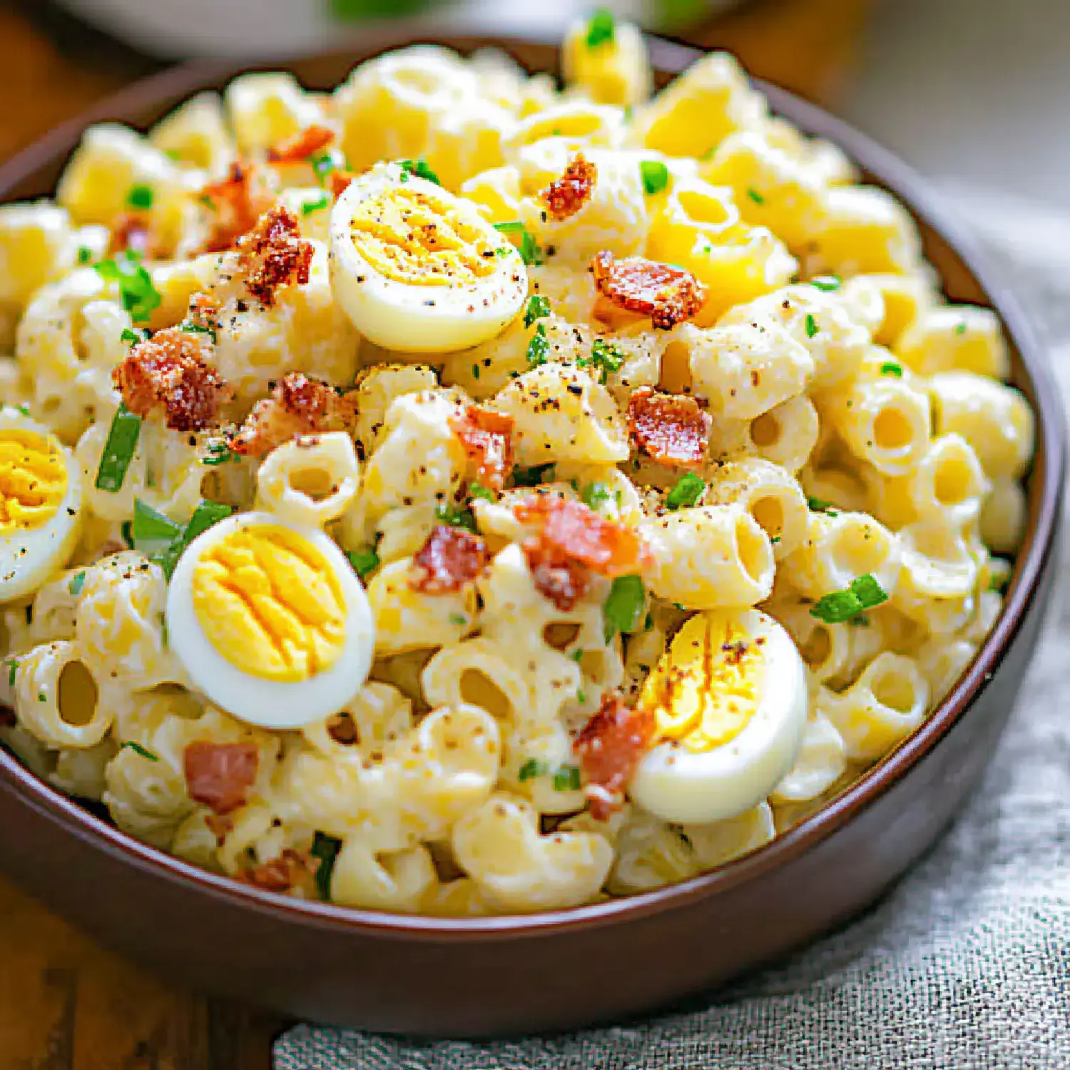 A bowl of creamy pasta salad topped with sliced hard-boiled eggs, crispy bacon bits, and chopped green onions.