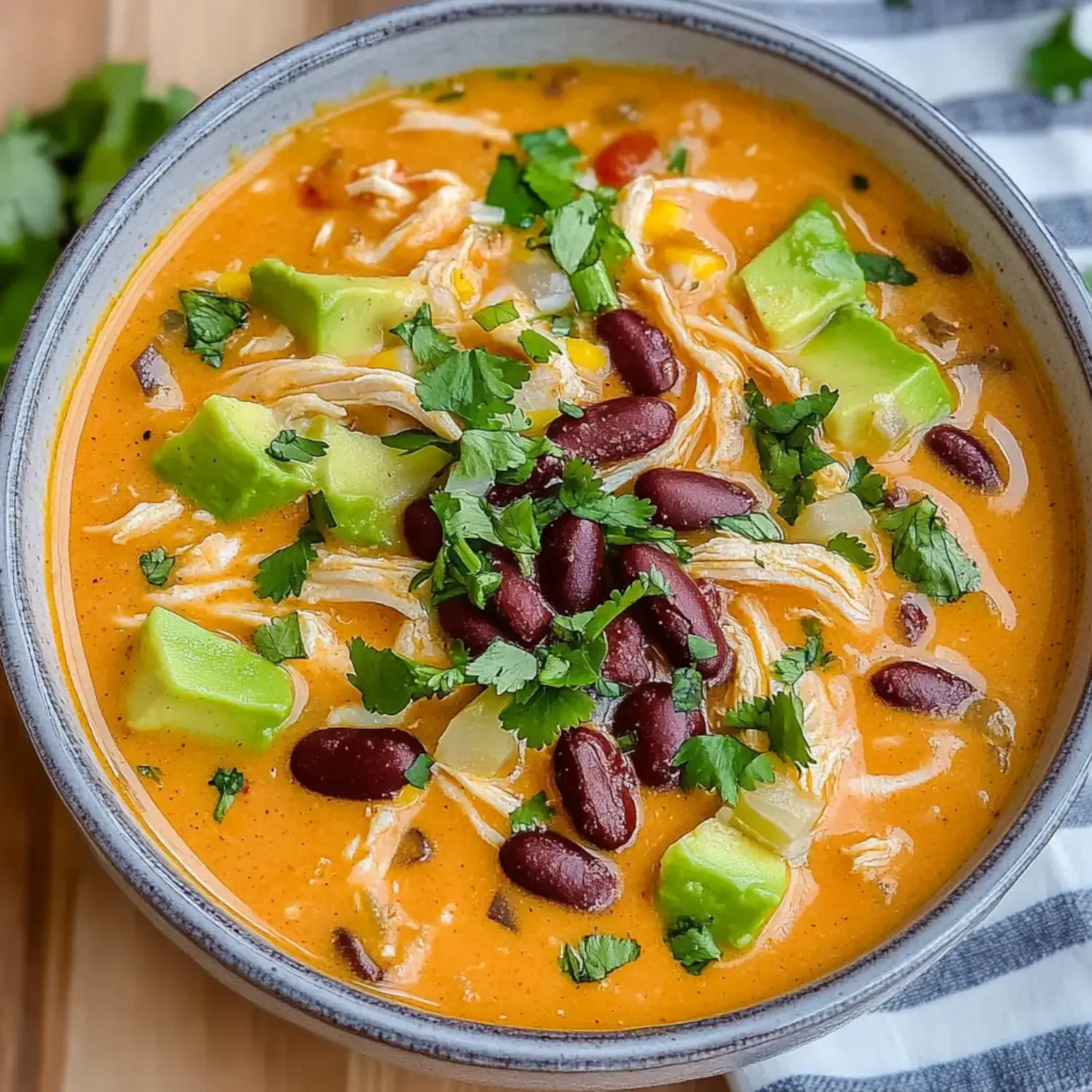 A bowl of creamy orange soup topped with shredded chicken, diced avocado, red kidney beans, corn, and fresh cilantro.