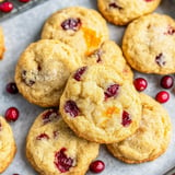 A close-up of freshly baked cookies studded with cranberries and orange peel, surrounded by red cranberries on a light surface.