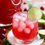 A glass of pink drink with ice and a lime wedge sits on a white coaster, surrounded by fresh limes and cherries.