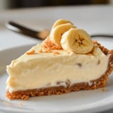 A slice of banana cream pie topped with banana slices and a sprinkle of crust crumbs on a white plate.