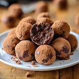 A plate of chocolate truffles dusted with cocoa powder, with one truffle cut in half to reveal its rich, dark filling.