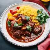 A plate of rich beef stew garnished with cilantro, served alongside yellow rice and colorful bell peppers.