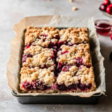 A freshly baked berry crumble dessert cut into squares, in a lined baking pan, with berries and jam nearby.