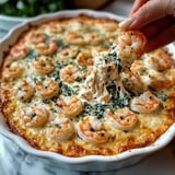 A hand is pulling a cheesy shrimp and spinach dip from a white serving dish, showcasing shrimp on top and melted cheese stretching from the dish.