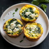 A plate of three baked spinach and cheese muffins is garnished with crumbled white cheese and sprinkled with black pepper.