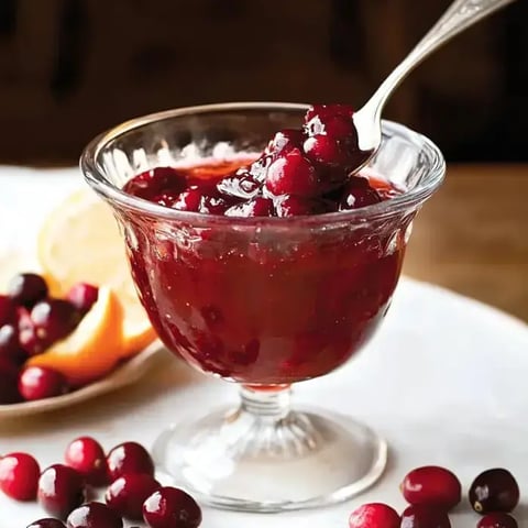 A glass dish filled with cranberry sauce, garnished with whole cranberries, and a spoon resting on the rim.